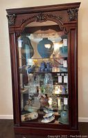 Front view of the dark wood curio cabinet showing decorative carved top and shelves filled with various ceramic and glass items