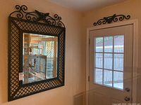 Full view of the beveled mirror with black lattice metal frame and decorative top scroll with bird design, hanging on wall near door.