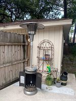 Full view of the propane outdoor heater positioned on concrete against a wooden fence and garden backdrop, showing the full height and details of the heater including base, pole, and top reflector.