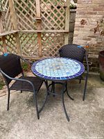 Outdoor patio set with ceramic mosaic round table and two black woven chairs on metal frames placed on concrete surface near wooden lattice fencing and brick wall.