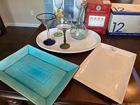 Photo showing three trays stacked: turquoise stoneware reactive glaze tray in front, white oval porcelain tray behind, and a smaller white rectangular porcelain tray underneath, with several glasses on top of the trays.