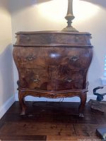 Full front angle of dark wood side table with three drawers, showing curved shape and ornate metal drawer handles.