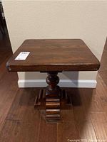 Front view of the small solid wood table showing the square top, turned central pedestal column, and wide base with feet on wooden floor against a wall.