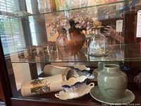 Photo showing the contents inside a wooden and glass display cabinet including the pottery gravy boat, signed pottery vase and plate, ceramic vases, cactus motif shot glasses, and Patron decanter.