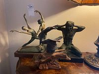 Three dancing statues on a wooden surface, including a ballet couple, a seated male figure, and a decorative abstract piece.