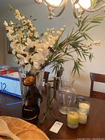 Wide view showing tall cut glass vase with faux florals, metal cat wine bottle holder, clear vase, candle holders and candles on table