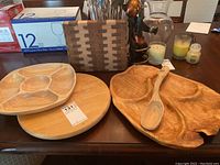 Photo showing three wooden serving pieces including a chip and dip tray, lazy Susan, and serving platter with spoon, plus boxed drinking glasses and decorative items in background.