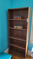 Full view of tall laminate composite bookcase against blue wall showing all five shelves and some books temporarily placed for staging.