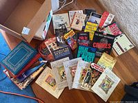 Multi-book assortment showing Marvel Masterworks comics with vintage covers, true crime, history, and other paperback books spread on floor with a cardboard box.