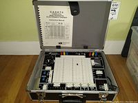 Open aluminum carrying case containing trainer modules, central solderless breadboard, and instruction manual attached to lid
