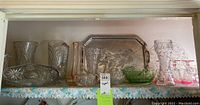 Wide shelf view showing clear and green cut glass items including tall vases, a pitcher, a green footed bowl, and a metal serving tray.