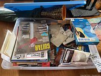 Overhead view of a plastic bin filled with assorted guitar magazines, instructional books on guitar building, guitar strings, and a disassembled plastic Didgeridoo with cloth carrying bag beside it.
