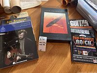 Photo shows a mix of guitar instruction and music books, including Joe Satriani Guitar Secrets, Led Zeppelin discography book with distinct cover art, and Guitar Legends DVD.