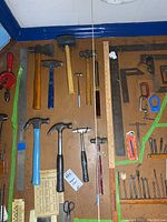 Photo showing various hammers including claw hammers, ball peen hammers, a rubber mallet, metal clamps, measuring sticks, wooden rulers, metal squares and other hand tools arranged on a pegboard.