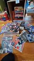 Photo showing New England Patriots magazines with players in uniform featured on covers, including Tom Brady and others, several NFL-related DVDs in plastic cases, and books partially visible on shelves in background.