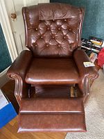 Front view of reclining armchair showing brown leather-feel upholstery, button tufting, padded arms, and extended footrest.