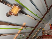 Two fishing rods mounted horizontally on a wall. One rod has a large vintage-style Penn reel with yellow line and wooden handle with decorative wraps. The other rod has a foam handle with a red Penn reel. Additional fishing gear storage and tools visible nearby.