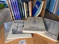 Books and printed ship model plans, highlighting detailed ship line drawings of three historical ship models laid out on table with boxed books in background
