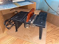 Photo of Dremel table saw with black metal body and reddish plastic blade guard resting on cardboard in original box, showing power cord and switch.