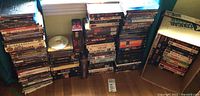 Stack of VHS and DVDs in various sizes and cases on floor near window, showing large collection of movies