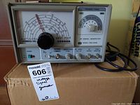 Front view showing the analog meter, control knobs, frequency range selector, power switch, cord connecting at rear, and the original cardboard box beneath unit.