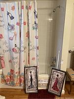 View of two Kirklands framed prints leaning against the bathtub, shower curtain displaying high heels and perfume bottles with flower and text designs.