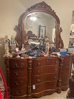 Front view of the wood dresser with ornate carved detailing, multiple drawers, and a large mirror mounted on top, with various small items on top of dresser