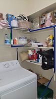 Wide view of laundry area showing various household items on wire shelves including cleaning supplies, paper towels, bottles, storage containers, and a black bag.