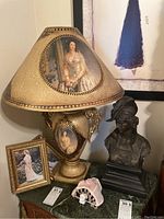 Photo showing 28-inch resin table lamp with matching shade depicting Victorian-style portraits, 18-inch dark resin bust on black base, framed 5x7 print in gold frame, and small decorative miniature bonnet.
