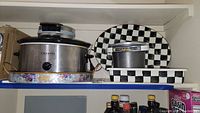 Wide shot showing Crockpot, floral edge ceramic bakeware, Presto FryDaddy deep fryer, and two MacKenzie-Childs black and white checkered serving trays stacked behind.