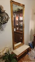 Front angled view of tall gold-framed wall mirror and small bench with faux marble top and gold legs.