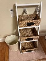 Photo showing front view of white wooden organizer rack with three woven wicker baskets on each shelf and beige trash can to the side on a light-colored bath mat.