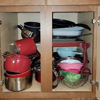 View of the red and stainless steel pots and pans, assorted bowls, baking pans, and utensils inside two cabinet doors.