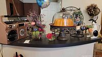 View of reproduction radio on counter next to faux floral arrangement, bunny pitcher, cake stand with faux Bundt cake under glass dome, and carrot-shaped wine bottle holder.