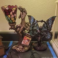 Two Dale Tiffany stained glass lamps shown side-by-side on countertop; one with floral stained glass shade and bronze female figure base, other is Lady Butterfly lamp with blue stained glass wings.