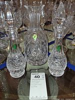 Three Waterford crystal pieces: two cruets with stoppers and one bottle without stopper, placed on glass table with additional glassware in background.