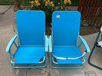 Two blue metal framed beach chairs with blue fabric seats and backs, placed side by side on a concrete surface near yellow flowers.