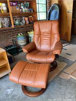Full view of recliner and ottoman in tan leather on wooden round swivel bases, set in garage