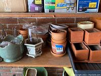 Image shows a variety of small flower pots including terra-cotta, square pots, and decorative ceramic pots arranged on a wooden shelf.