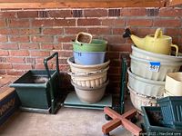 Photo showing stacked beige ceramic pots of various sizes, green and blue plastic pots, green rolling plant stand, and padded gardening kneeler wooden legs.
