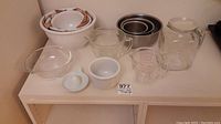 Photo showing all the items included in the set on a white shelf: nested white ceramic mixing bowls, nested metal mixing bowls, clear glass pitcher, measuring cup, reamer, and glass dish.