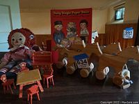 Raggedy Andy doll with red yarn hair and red checkered outfit. Wooden red dollhouse furniture including a table, chairs, a small bed or crib. Four wooden pull horse toys on wheels. Dolly Dingle Paper Dolls box propped against the wall.