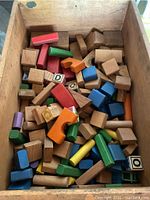 Wooden building blocks in various colors and shapes inside a wooden box, showing letters and solid colors.