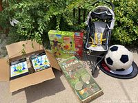 Wide shot showing all items including two boxed packages of soft tip darts, wobble deck game, tennis set box, doll stroller, plush soccer ball on black plastic base, soft frisbee, with leafy shrub background