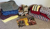 Seven large stacked afghans in various patterns and colors next to basket with crafting supplies and vintage crochet magazines in front.