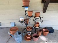 Wide view showing three black metal plant stands with curled decorative tiers holding multiple terracotta and plastic plant pots. Several terracotta pots and a galvanized metal watering can are placed on the concrete floor in front.