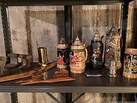 Overview of lot items on shelf in basement including five German beer steins, two wooden railroad track bookends, wooden boomerang, and wooden duck decor.