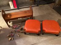 Full view of wooden bench, two foot stools with orange cushions, and metal flower decor on floor