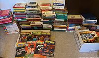 Large grouped stack of various books including multiple hardcover and softcover books, two boxes with smaller books and magazines, and Star Trek magazines laid out in front showing colorful covers.
