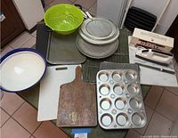 Full view of cookie baking items on table including cookie sheet, muffin tins, cooling racks, ceramic bowl, cutting boards, colanders, and electric cutting utensil in box.